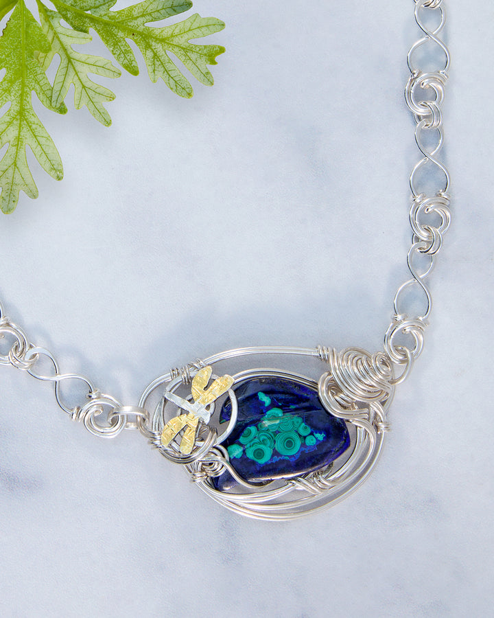 Silver necklace with a blue pendant featuring a dragonfly design on a light gray background.