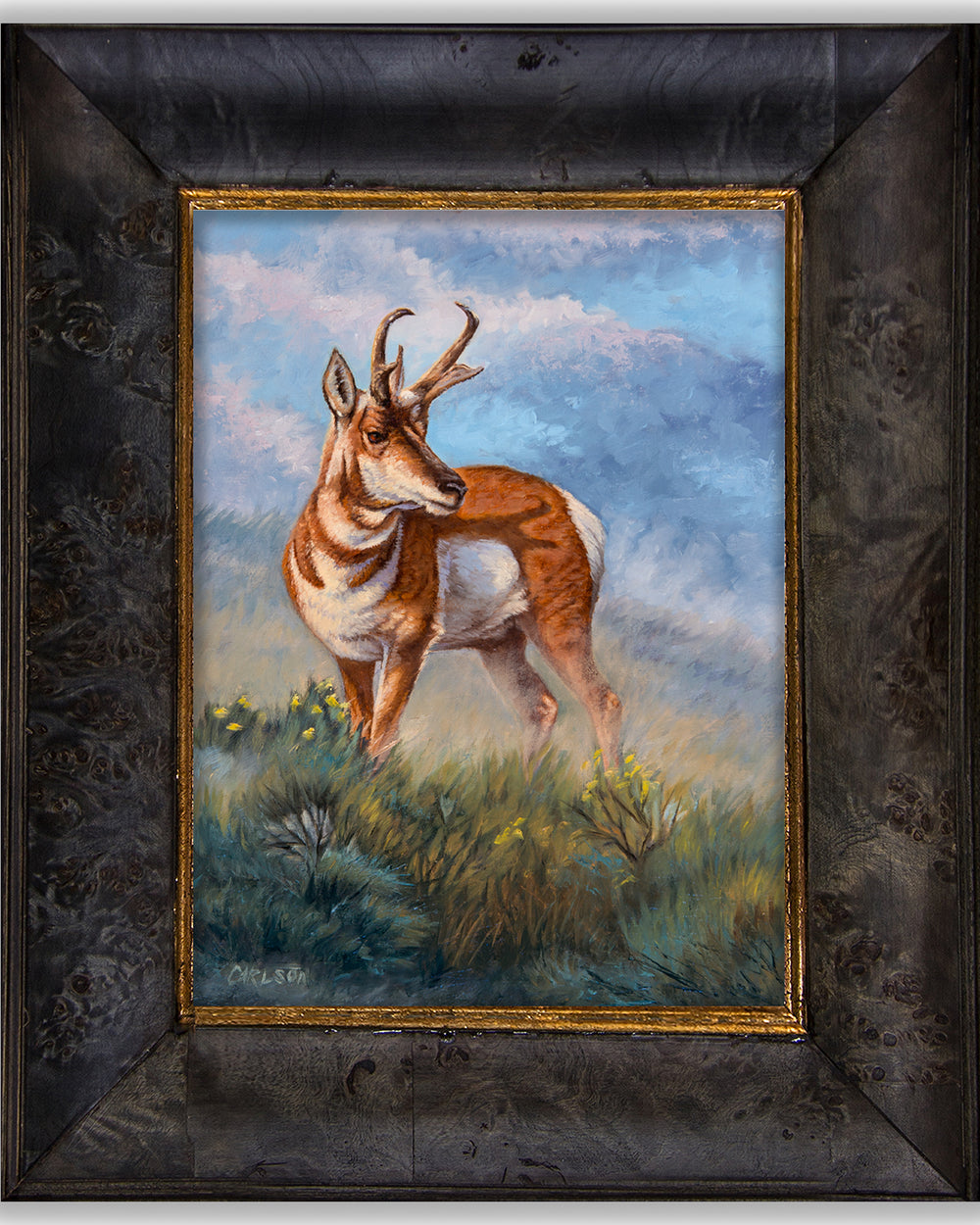 A framed painting of a pronghorn standing in a grassy field under a blue sky, with a dark textured frame and gold inner border.
