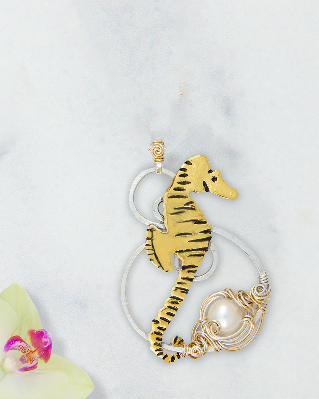 A seahorse-shaped pendant with black detailing, wrapped in gold- filled wire with a white genuine pearl accent, displayed on a light marble background with a small orchid flower in the corner.