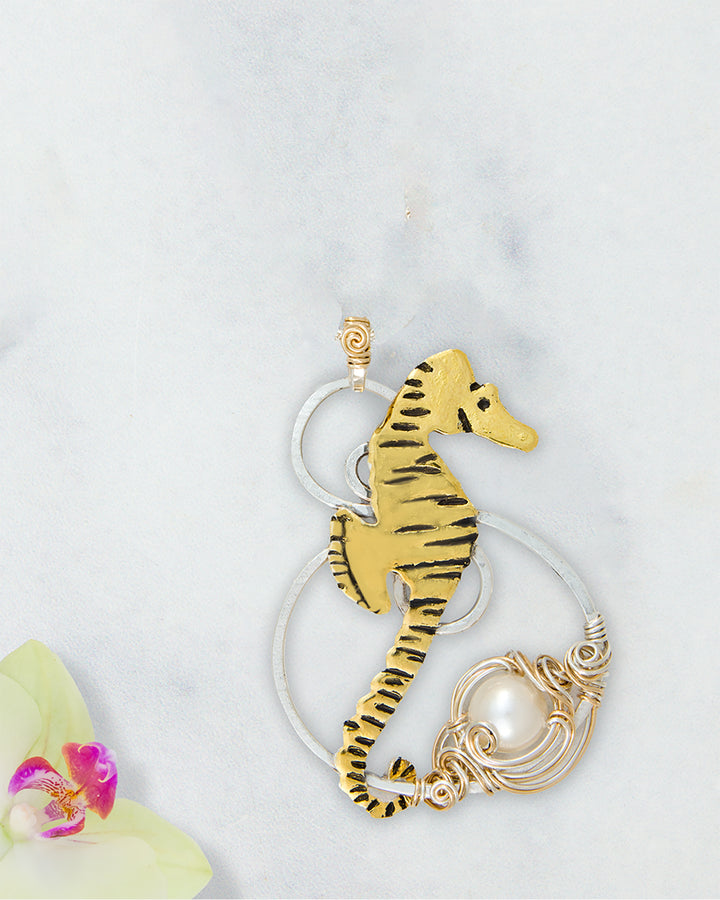 A seahorse-shaped pendant with black detailing, wrapped in gold- filled wire with a white genuine pearl accent, displayed on a light marble background with a small orchid flower in the corner.
