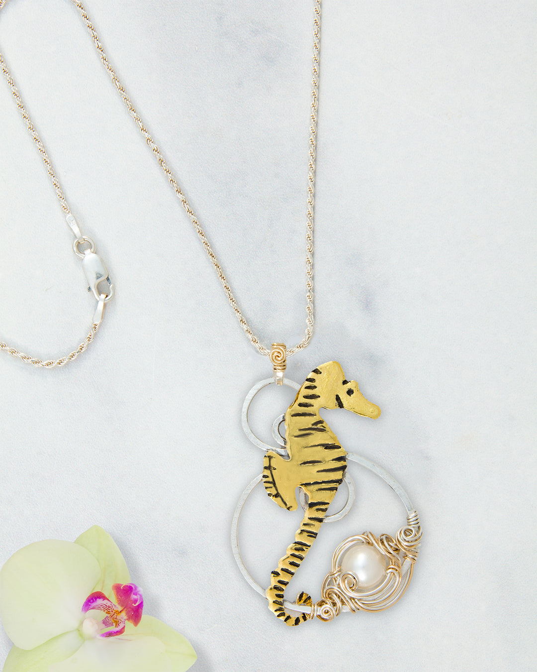 A gold and silver seahorse pendant necklace with a white pearl accent, displayed on a light marble background next to a white orchid flower.