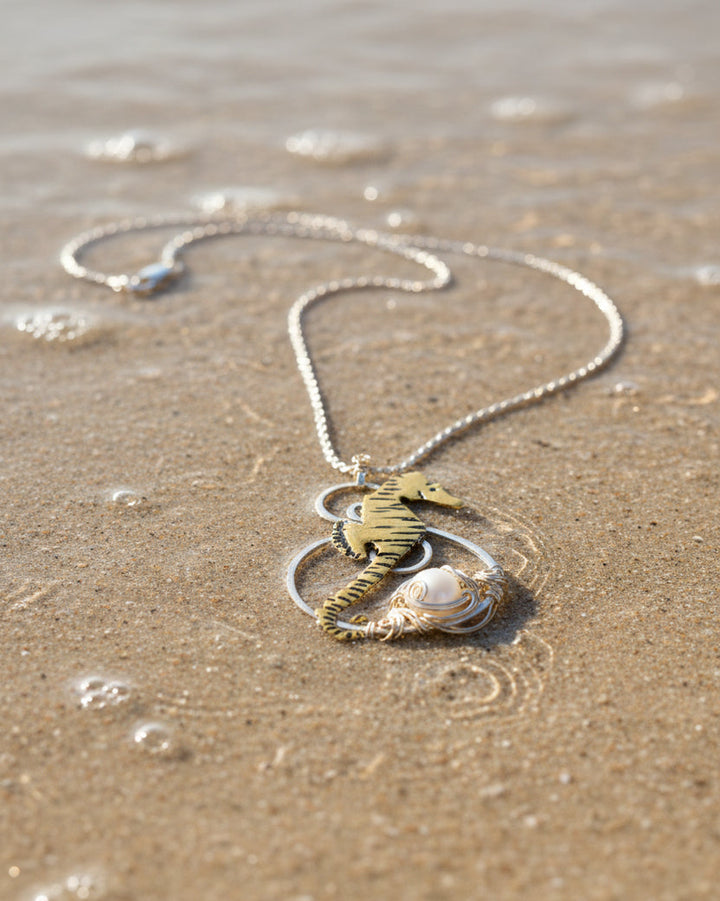 A gold and silver seahorse pendant necklace with a white pearl accent, displayed on a sandy beach