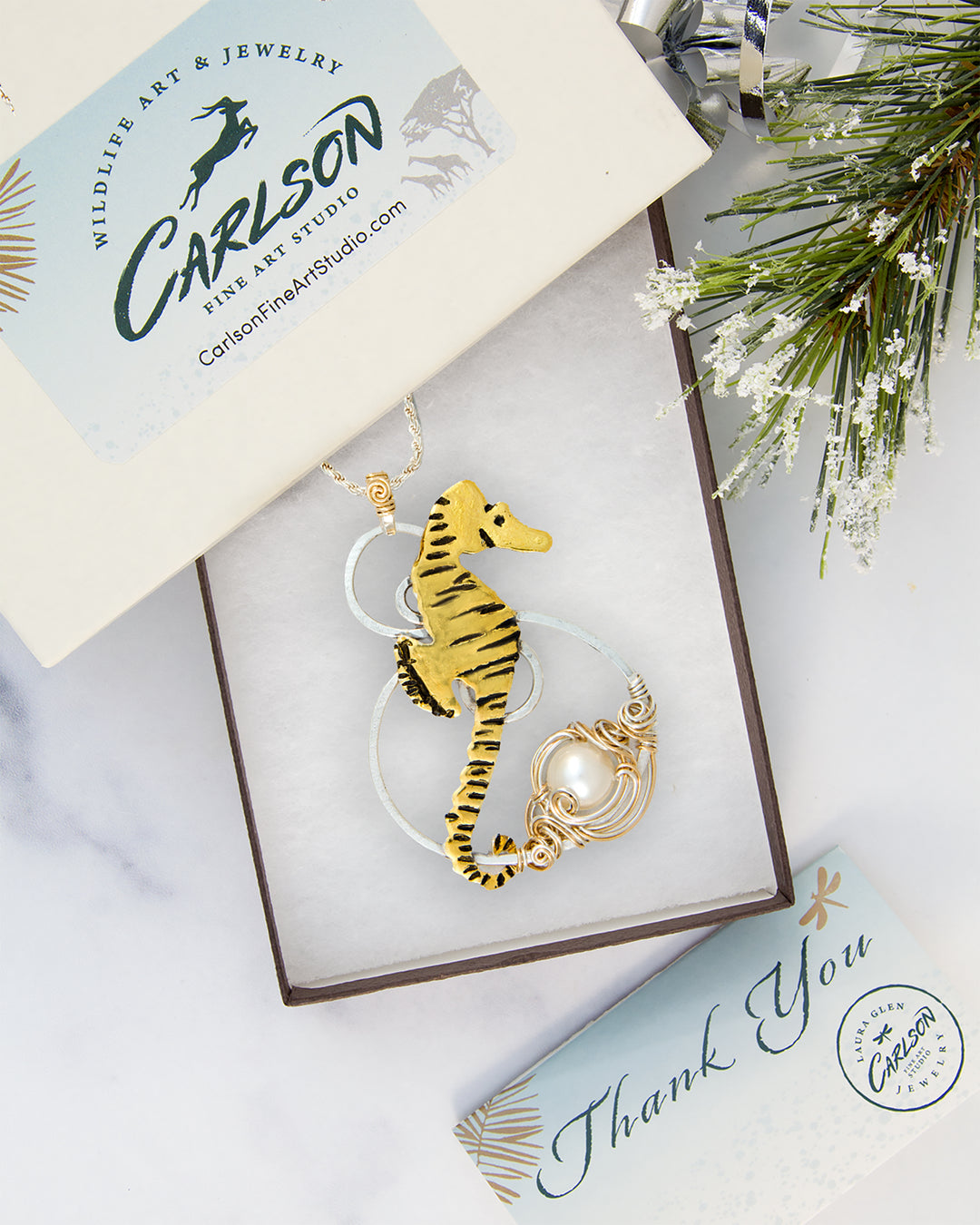 A gold seahorse pendant necklace with a white pearl accent, presented in an open gift box. The box lid features the Carlson Fine Art Studio logo, and the scene is styled with a pine branch and a thank you card on a white marble background.