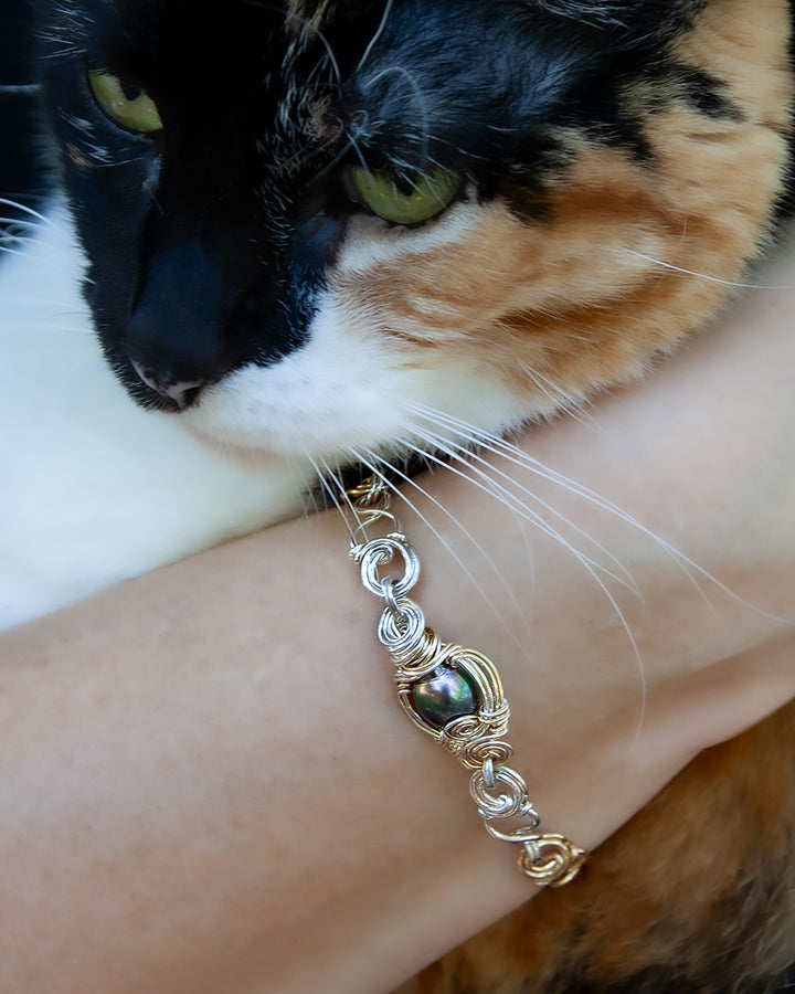 A close-up lifestyle shot of a handmade sterling silver and gold chain bracelet with an iridescent black Tahitian pearl, worn on a person's wrist with a calico cat resting on their shoulder.