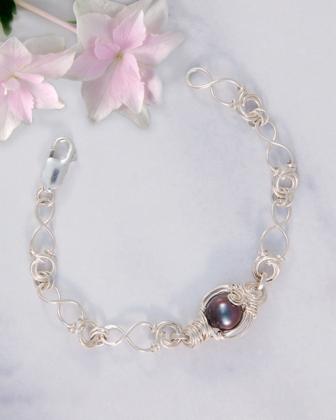 A silver chain bracelet with a Tahitian  pearl in a decorative setting, styled on a light marble surface with pink flowers in the corner.