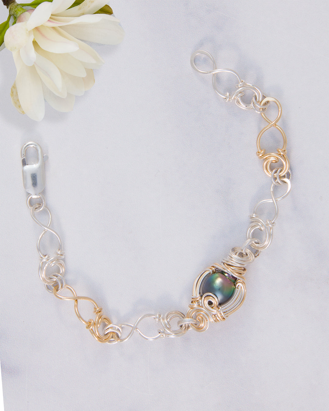 A handmade link bracelet featuring a central iridescent black Tahitian pearl set in gold-filled wire, with alternating silver- and gold-filled metal links, displayed on a light gray marble surface with a white flower in the corner.