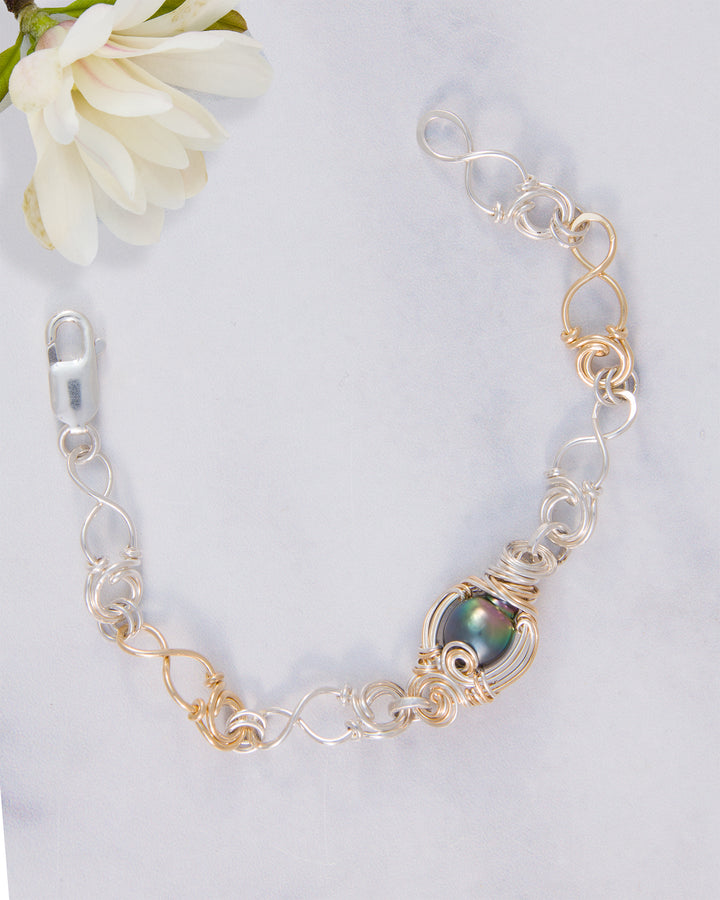 A handmade link bracelet featuring a central iridescent black Tahitian pearl set in gold-filled wire, with alternating silver- and gold-filled metal links, displayed on a light gray marble surface with a white flower in the corner.