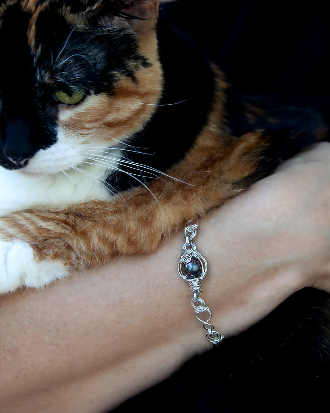 A close-up lifestyle shot of a silver wire-wrapped bracelet with a Tahitian pearl, worn on a person's wrist with a calico cat resting on their shoulder.