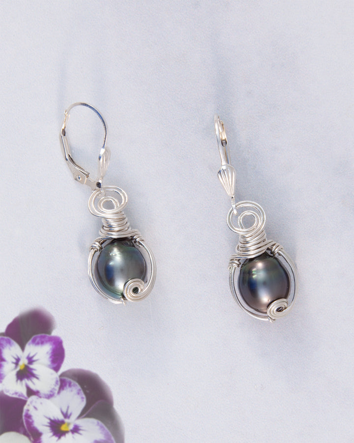 A pair of silver leverback earrings featuring dark, iridescent Tahitain pearls wrapped in delicate silver wire, displayed on a light gray background with purple flowers in the corner.