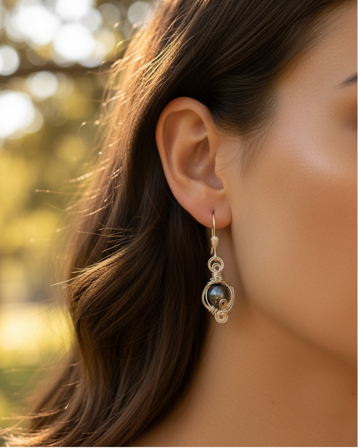 A close-up side profile of a woman wearing a delicate silver dangle earring with a dark iridescent pearl set against a warm, sunlit, and blurred outdoor background.