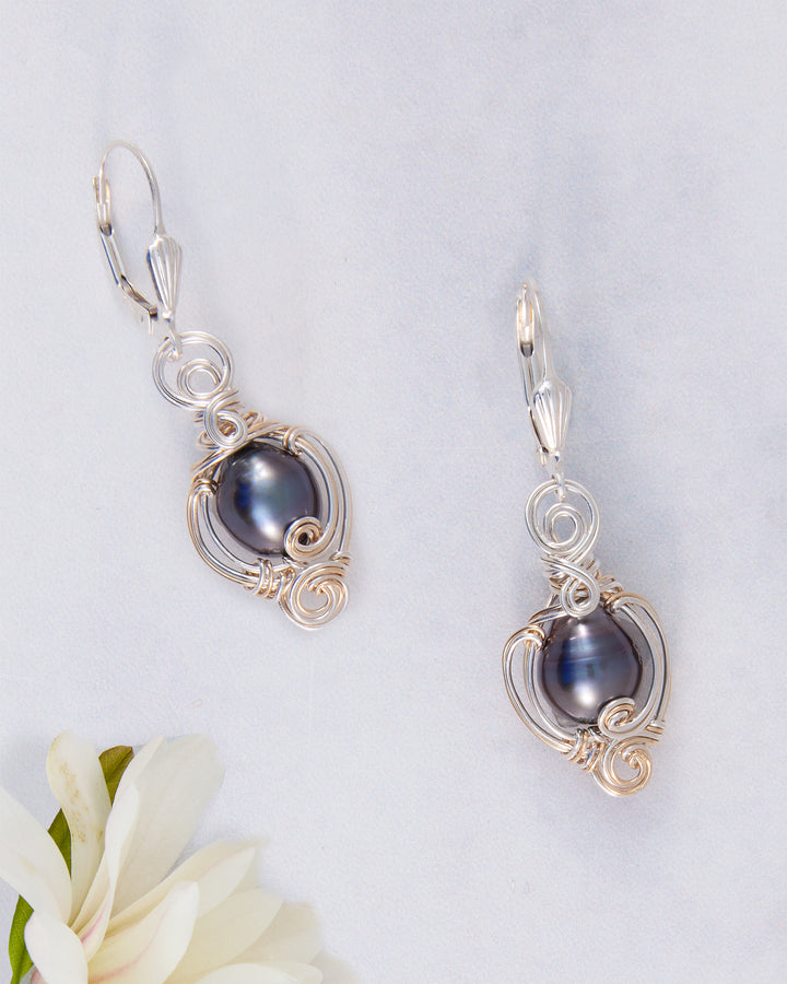 A pair of silver leverback earrings featuring oval dark blue-gray Tahitian pearls wrapped in delicate, ornate silver wire, displayed on a light textured background with a white flower in the corner.