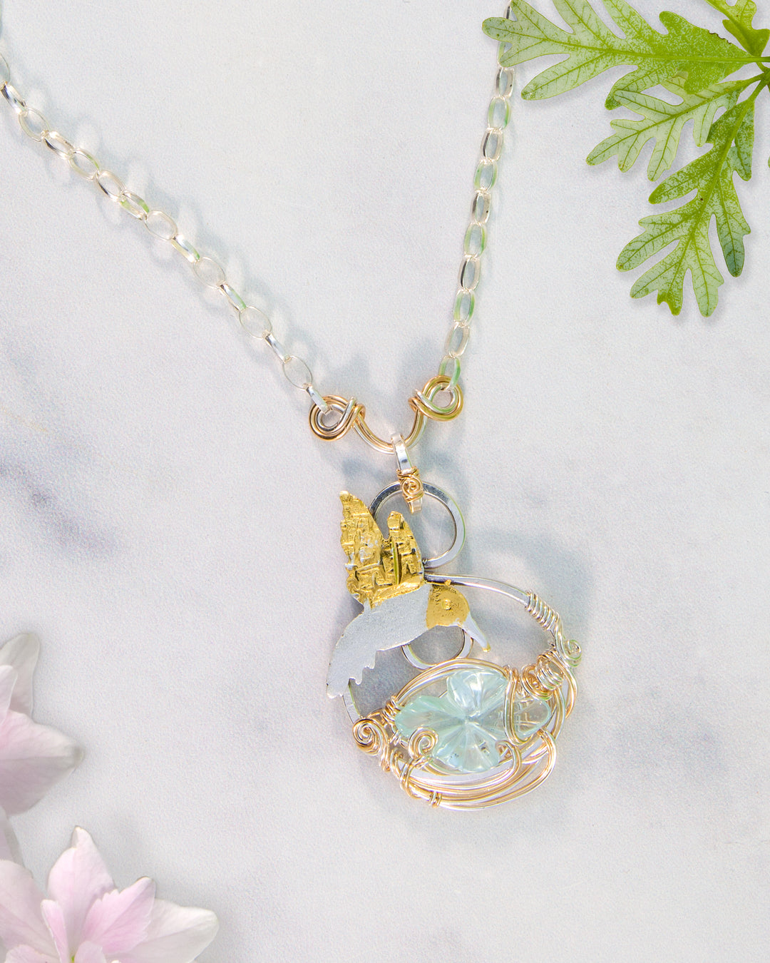 Necklace with a pendant featuring an aquamarine flower gemstone and hummingbird and silver and gold accents on a light background.