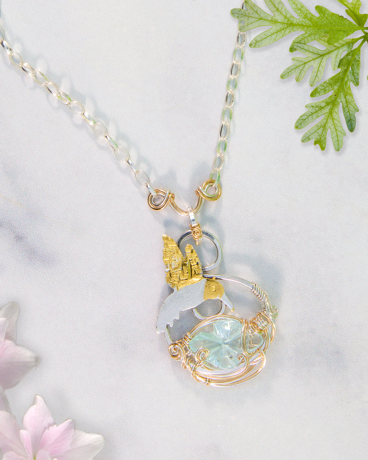 Necklace with a pendant featuring an aquamarine flower gemstone and hummingbird and silver and gold accents on a light background.