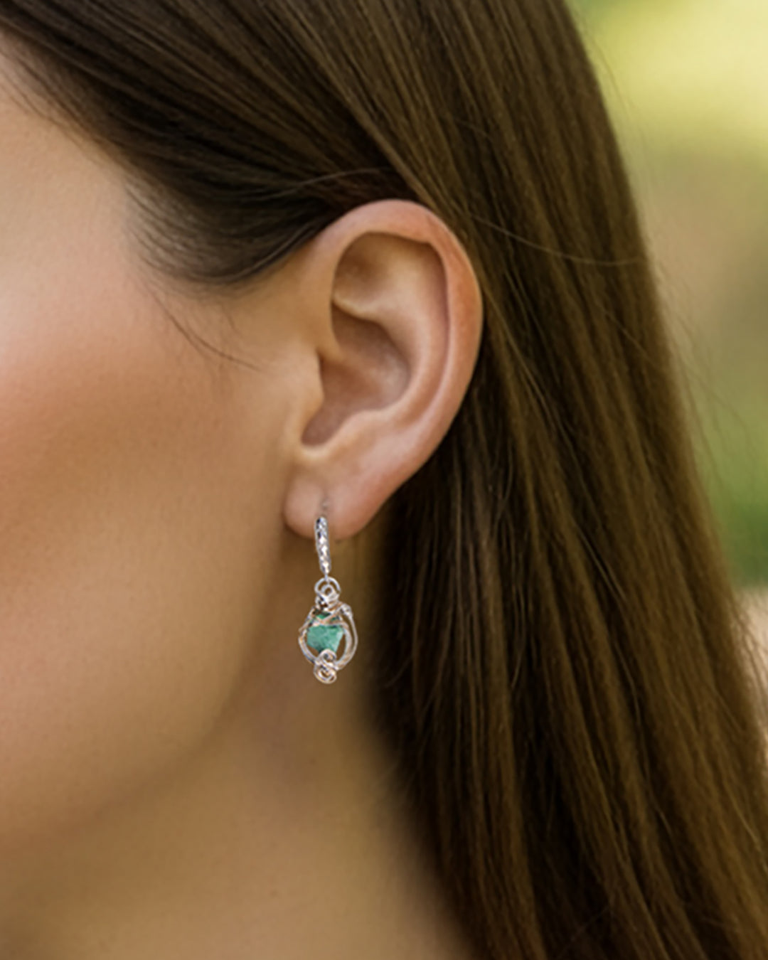 Close-up of a woman's ear wearing a silver earring with a green gemstone, blurred background