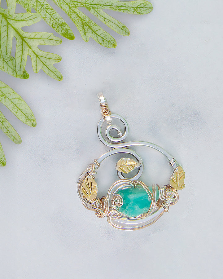 Silver and gold pendant with a green gemstone on a light gray background with green leaves.